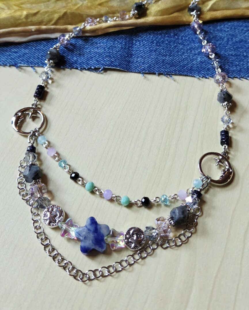 Close-up of the handmade Sodalite Star & Moon Necklace from Indigo Dusk, featuring blue goldstone, steel-blue and lavender crystals, a sodalite star, silver moon and star charms, and draped silver chain.