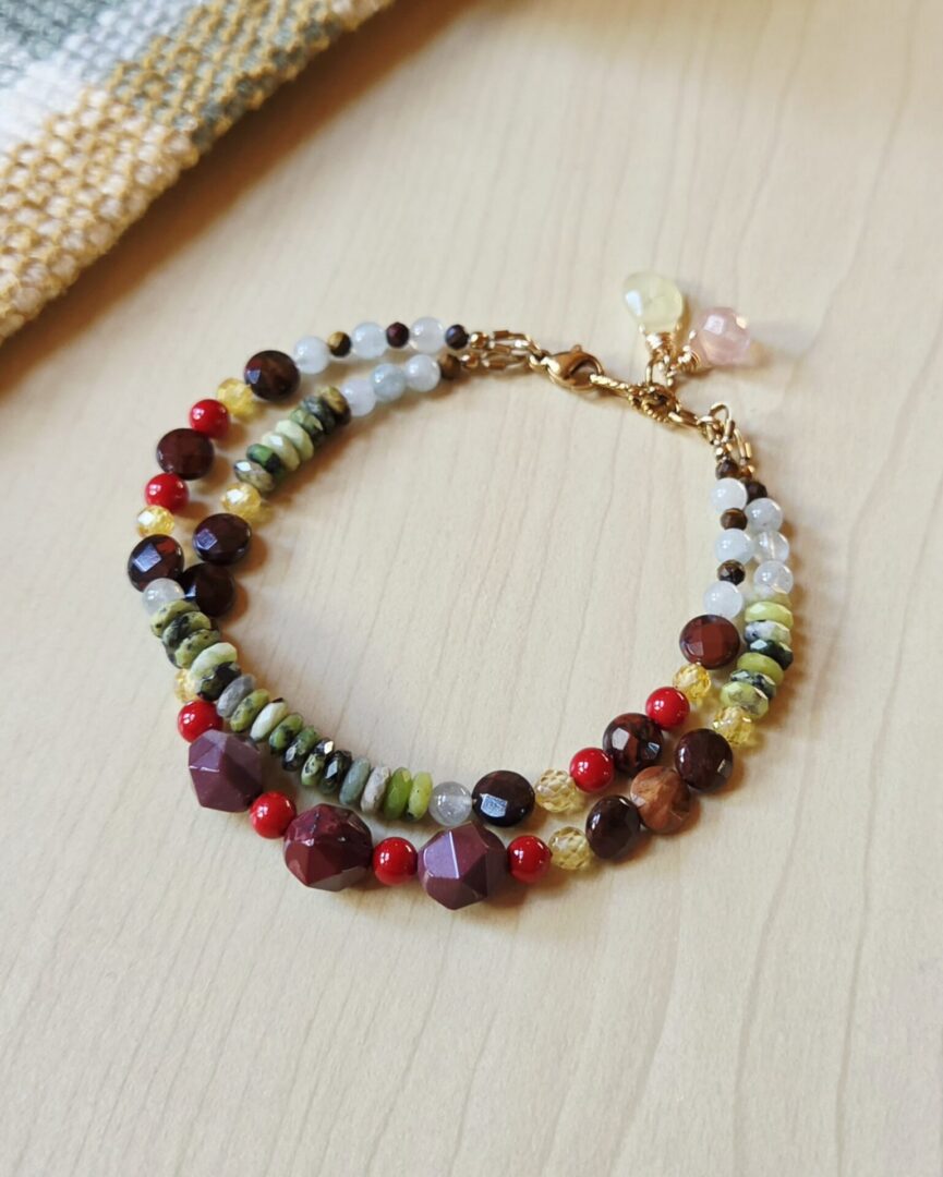 Handmade Mookaite and serpentine gemstone bracelet featuring earthy green heishi beads, burgundy and brown gemstone beads, bright red coral accents, golden zircon beads, and quartz teardrop charms with gold-tone chain.