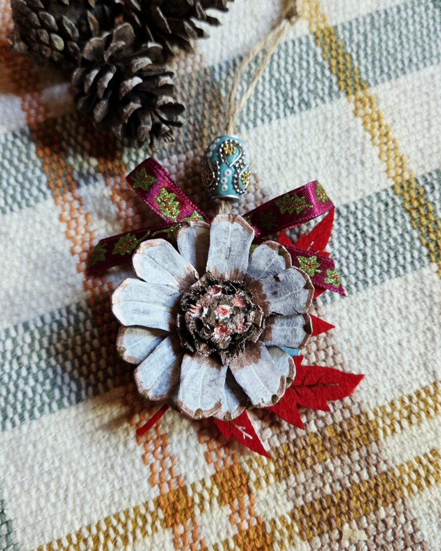 Handmade pinecone flower ornament painted white, copper, and salmon, featuring a red felt maple leaf, purple leaf-patterned ribbon, turquoise decorative bead, and rustic sisal hanger.