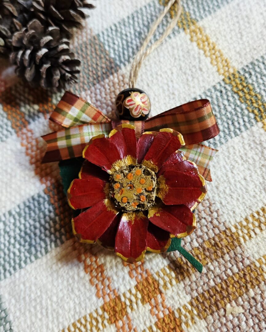 Handmade red pinecone flower ornament with gold and orange dotted center, green felt leaf, copper green and cream plaid ribbon, and floral-painted lacquer bead.
