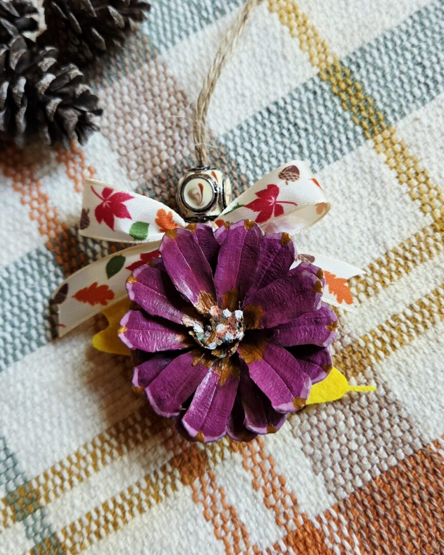 Handmade purple pinecone flower ornament with lavender and copper accents, yellow oak leaf, cream leaf-and-acorn ribbon, enamel-painted metal bead, and sisal twine hanger.